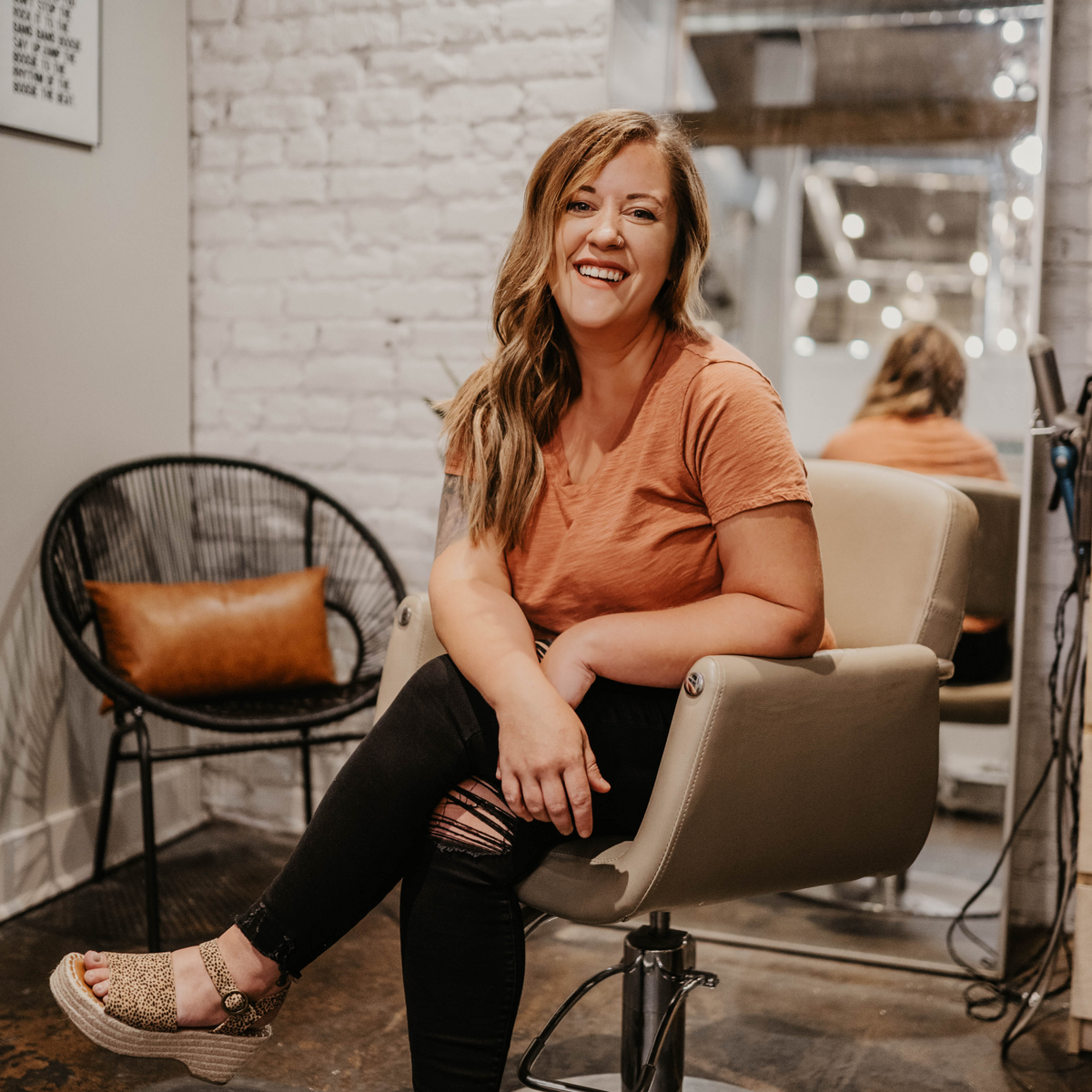 Ashleigh Stephens sitting in a salon chair smiling in front of a brick wall.